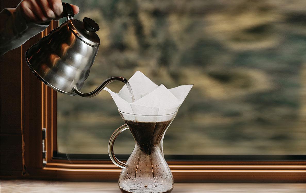 Ricco Coffee – Pour-Over Brewing Guide Banner Gooseneck kettle pouring hot water over a paper filter in a glass pour-over carafe, representing Ricco Coffee’s step-by-step brewing guides for making the perfect specialty coffee at home