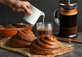 French-Press-Coffee-and-Cinnamon-Rolls A hand pouring cream into a glass of coffee, with a French press and freshly baked cinnamon rolls in the background, embodying the delightful experience of enjoying a rich and complex cup of French press coffee paired with sweet pastries.