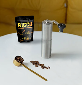 Perfect-Grind-Ricco-Coffee-Setup A pack of Ricco Coffee Company beans alongside a manual coffee grinder and a wooden scoop with beans, ready for grinding to the ideal coarse size for a French press, on a white table against a soft yellow background.