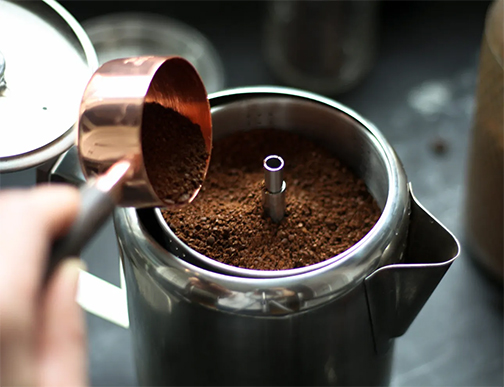 A person's hand is captured pouring finely ground coffee from a copper scoop into the stainless steel basket of a stovetop percolator, preparing for a traditional brew. The focus on the grounds and basket details highlights the initial step in achieving the full-flavored coffee that stovetop percolators are renowned for. This moment is a key part of the brewing ritual, setting the stage for a rich and aromatic coffee experience.