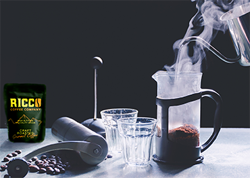 Ricco-Coffee-Company-Bag-With-French-Press-Grinder Steam rises from a French press beside a bag of Ricco Coffee Company beans and a stainless steel burr grinder, capturing the ideal preparation for a richly flavored brew.