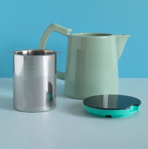 The disassembled parts of a Sowden SoftBrew coffee maker, including the stainless steel filter and teal lid, laid out beside the ceramic pot on a blue background.