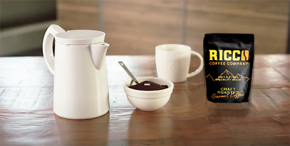 A Sowden SoftBrew coffee maker elegantly placed next to a mug and a Ricco Coffee Company bag of specialty grade premium gourmet coffee on a reflective wooden table, inviting a satisfying coffee experience.