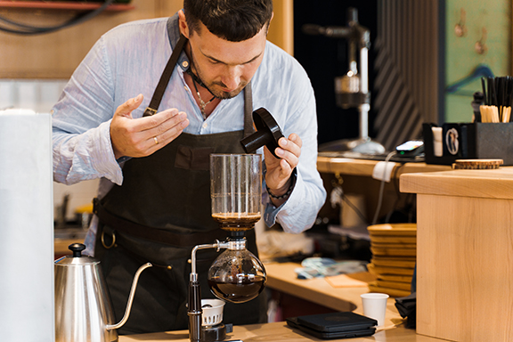 barista-preparing-siphon-coffee-maker.jpg A focused barista in an apron carefully setting up a vacuum siphon coffee maker, exemplifying the precision and expertise required in the siphon brewing process.