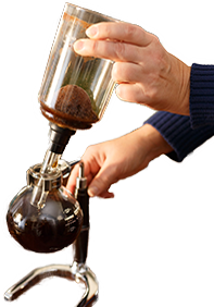 A person's hands demonstrating the initial step in a vacuum siphon coffee maker guide, attaching the upper chamber filled with coffee grounds to the lower chamber with brewed coffee, on a wooden table.