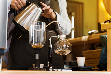 A barista carefully pouring hot water into a siphon coffee maker, an intricate step to prevent over or under extraction, ensuring a perfectly balanced cup of coffee.
