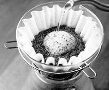 Black and white image capturing the initial pour of hot water over coffee grounds in a paper filter, starting the blooming process during pour over brewing to enhance flavor extraction.